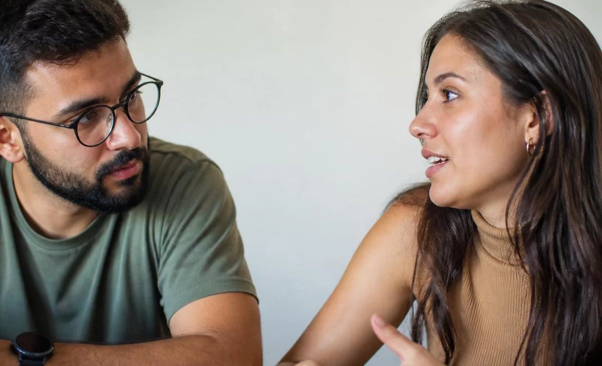 Três participantes conversam ao redor de uma mesa comprida numa sala clara de janelas altas